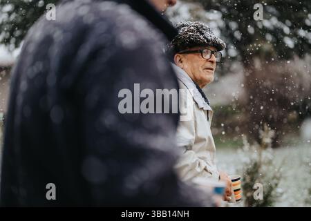 Homme âgé par temps neigeux tenant une tasse rayée et réfléchissant à l'extérieur Banque D'Images