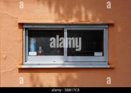 Fenêtre rectangulaire en verre figuré dans un mur orange texturé en stuc. Les arbres environnants créent une ombre de type yin-yang, créant une tranquillité zen. Banque D'Images