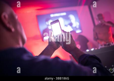 Rome, italie 4 mai 2025 : spectateur enregistrant un dj set avec smartphone dans une discothèque avec écran blanc vierge et éclairage rouge Banque D'Images