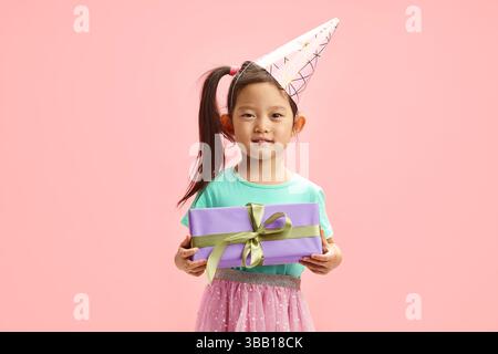 Charmante fille asiatique d'enfant autour de 5-6 ans dans le chapeau de fête, portant dans des vêtements de fête, tenant la boîte-cadeau sur fond isolé rose Banque D'Images