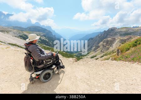 L'homme handicapé utilisant un fauteuil roulant observe des paysages de montagne magnifiques dans les Dolomites. La lumière du soleil illumine le paysage pendant qu'il prend dans le Banque D'Images