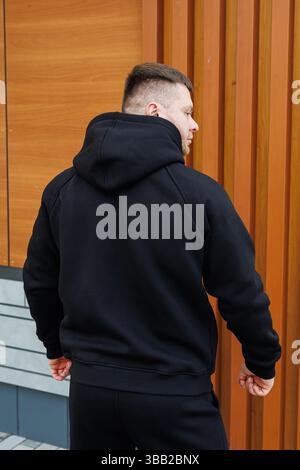 Un jeune homme vêtu d'un élégant sweat à capuche noir se tient debout, le dos face à l'appareil photo, affichant fièrement son style de mode urbaine contre un style moderne Banque D'Images