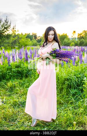 Une femme enceinte se tient debout dans un champ de fleurs violettes. Elle porte une robe rose et tient un bouquet de fleurs. La scène est sereine et paisible Banque D'Images