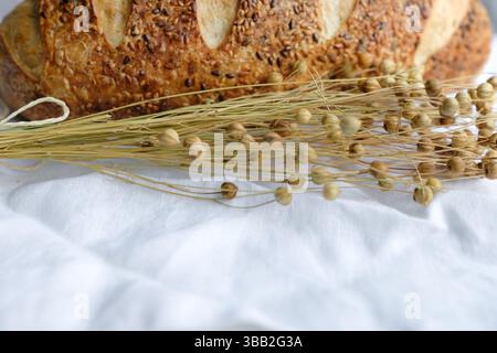Pain saupoudré de graines de lin et de sésame sur une nappe de lin blanc. Gousses de graines de lin. Pain au levain. Banque D'Images