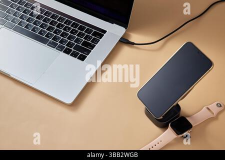 Ordinateur portable avec montre intelligente et smartphone se chargeant sur chargeur sans fil sur table beige, appareil de travail moderne Banque D'Images
