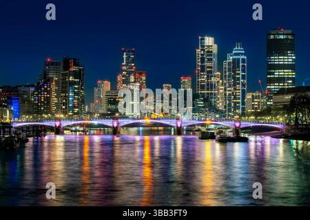 Horizon moderne de Londres la nuit avec des lumières colorées reflétant la Tamise et un pont illuminé au premier plan. Banque D'Images