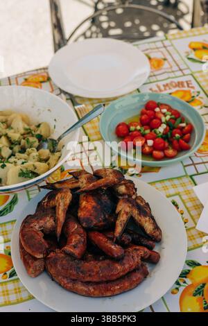 De délicieuses viandes grillées accompagnent une salade fraîche et des pâtes dans un cadre extérieur animé sous la chaleur du soleil. Banque D'Images