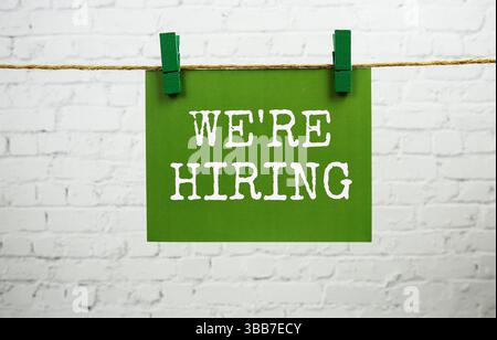 Nous embauchons du texte sur la carte de papier vert accroché avec de la corde sur fond de brique blanche Banque D'Images