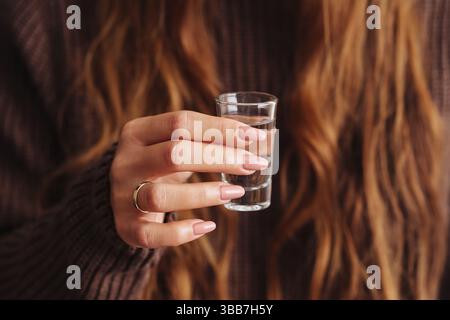 Gros plan d'une main de femme tenant un tir de vodka. Verre transparent avec de l'alcool. Manucure élégante et bague en or. Pull marron en arrière-plan. Mode de vie de fête. Banque D'Images
