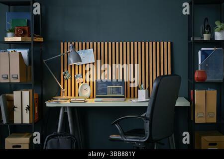 Installation moderne de bureau à domicile avec une toile de fond en bois, une lampe de bureau et un ordinateur portable sur une table blanche entourée d'étagères noires remplies de livres et de rangement Banque D'Images