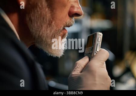 Homme senior examinant de près un petit appareil portable avec une expression ciblée. Porter des gants, indiquant sa participation à des travaux scientifiques ou techniques Banque D'Images