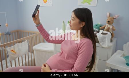 Femme enceinte prenant selfie dans une chambre confortable avec berceau de bébé, mettant en valeur ses longs cheveux foncés et portant une tenue rose, entourée de peluches et PA Banque D'Images