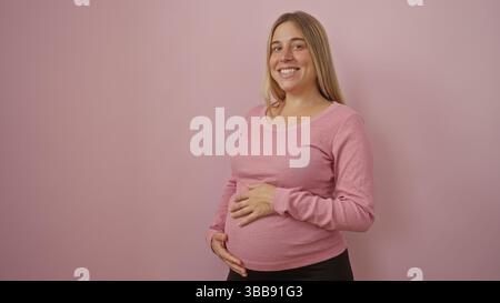 Femme enceinte aux cheveux blonds souriant et berçant son ventre se tient sur un fond rose doux, incarnant la joie et l'espérance dans un scen isolé Banque D'Images