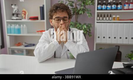Médecin portant des lunettes en manteau blanc dans le bureau de l'hôpital assis avec un ordinateur portable paraissant épuisé et stressé entouré d'équipement médical et de documentmen Banque D'Images