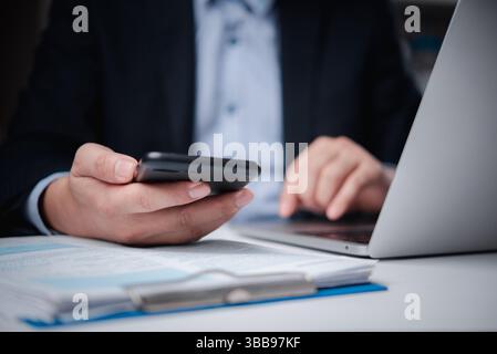 Gros plan d'un homme d'affaires tenant un smartphone et tapant sur un ordinateur portable, axé sur le travail professionnel. Banque D'Images