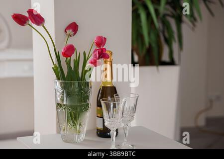 Une nature morte festive avec une bouteille de champagne avec deux verres aux côtés d'un bouquet vibrant de tulipes rouges dans un vase en cristal sur un surfa blanc Banque D'Images