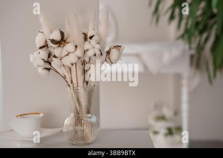 Un arrangement délicat d'herbe de pampa séchée et de tiges de coton dans un vase en verre côtelé se trouve sur une surface beige près d'une soucoupe blanche et d'une tasse à thé. Banque D'Images