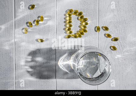 Capsules de vitamine d disposées en forme de lettre d sur une table en bois blanc à côté d'un verre d'eau, représentant l'importance de cette vitamine Banque D'Images