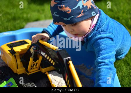 Garçon tout-petit joue avec la pelle à jouet dans un petit bac à sable dans une cour arrière. Enfant s'amusant dehors au printemps. Banque D'Images