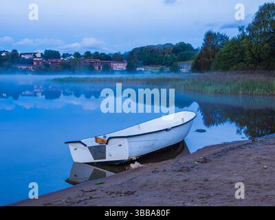 Un bateau solitaire repose sur la plage de sable d'un lac calme à Mölndal, en Suède. La brume matinale plane au-dessus de l'eau, créant une atmosphère sereine et tranquille Banque D'Images