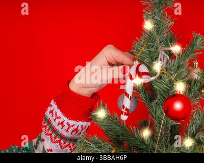 Image rapprochée de la main de femme caucasienne décorant l'arbre de Noël. Fond rouge. Dame en pull confortable mettant la canne de bonbons sur l'arbre. Banque D'Images