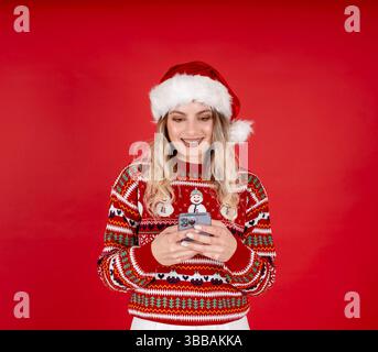 Portrait de la femme de Père Noël caucasienne mignonne excitée souriante à l'aide d'un téléphone portable. Fille 20s en chapeau de Père Noël rouge, pull de Noël. Saisie d'un message, isolé rouge Banque D'Images