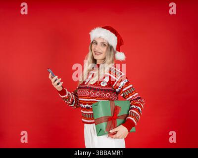 Femme caucasienne blonde heureuse tenant la boîte-cadeau cadeau cadeau de Noël enveloppant et la caméra regardant smartphone. Porter un pull chaud confortable, chapeau rouge. Banque D'Images