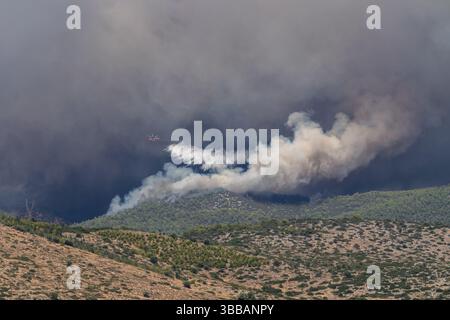 Un hélicoptère de lutte contre les incendies jette de l'eau au-dessus d'un feu de forêt. Une fumée dense monte au-dessus des arbres lors d’une intervention d’urgence suite à une catastrophe naturelle Banque D'Images