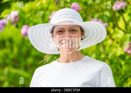 Une amicale femme d'âge moyen portant un chapeau pose sur un fond de nature et de fleurs Banque D'Images