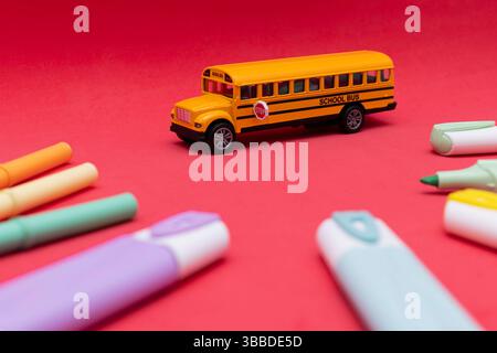 Autobus scolaire jouet placé sur une surface rouge, entouré de marqueurs de couleur et de marqueurs, représentant le thème de l'école Banque D'Images