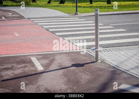 piste cyclable rouge traversant une traversée zèbre dans un cadre urbain moderne. La scène présente un poteau métallique projetant une ombre, un trottoir adjacent et un vert Banque D'Images
