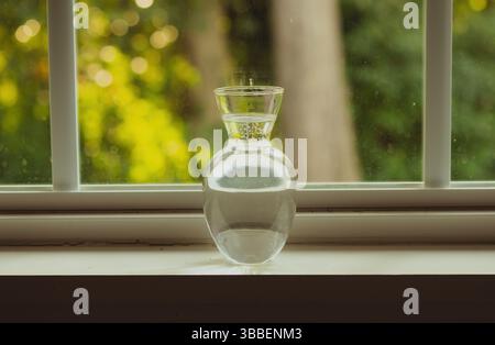 Un vase en verre simple et élégant rempli à mi-chemin d'eau se trouve sereinement sur un rebord de fenêtre. Banque D'Images
