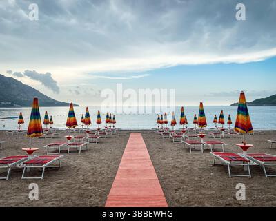 Ciel nuageux eaux calmes de la mer Méditerranée, rangées de transats vides et parasols sur la plage de sable au petit matin au Monténégro Banque D'Images