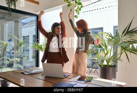 Deux femmes d'affaires heureuses célébrant le succès à la recherche d'un ordinateur portable dans un bureau vert. Banque D'Images