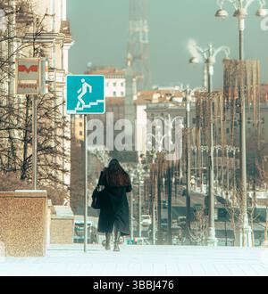 Scène de rue urbaine avec femme marchant près du panneau de métro et symbole d'escalier, capturé un jour froid dans le centre-ville. Banque D'Images