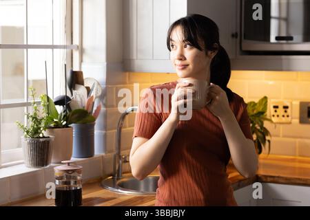 Femme asiatique debout tenant une tasse en céramique au comptoir de cuisine, avec des plantes en pot, espace de copie Banque D'Images