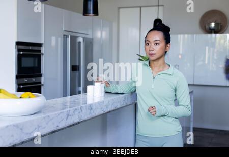 Femme asiatique debout tenant un verre d'eau à l'îlot de cuisine, avec des bouteilles de pilules blanches, espace de copie Banque D'Images