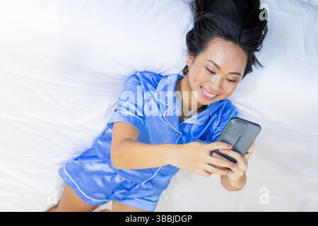 Femme asiatique couchée sur un lit couvert de draps dans la chambre tenant un smartphone noir et portant une bague en or Banque D'Images