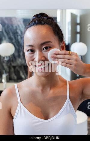 Femme asiatique appliquant un tampon de coton sur la joue dans la salle de bain, avec dosseret en marbre et miroir éclairé Banque D'Images