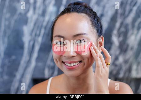 L'application de gel rose sous les yeux patches femme asiatique à la vanité murale en marbre, soulignant l'anneau d'or Banque D'Images