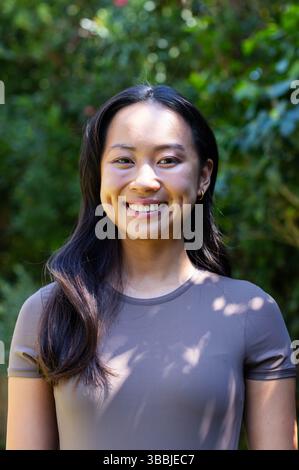 Femme asiatique souriante debout dans le jardin dans la lumière du soleil tapissée, portant des boucles d'oreilles cerceaux Banque D'Images