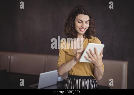 Femme souriante tapant tablette blanche à l'intérieur contre le mur texturé sombre, avec ordinateur portable argenté Banque D'Images