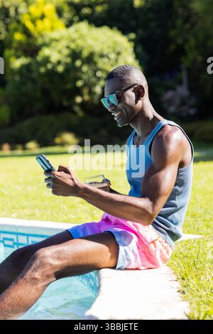 Homme afro-américain dans des troncs de bain plongeant les pieds dans la piscine arrière avec smartphone, espace copie Banque D'Images