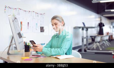 femme défilant smartphone au bureau dans le bureau ouvert, avec écran d'ordinateur et swatches Banque D'Images