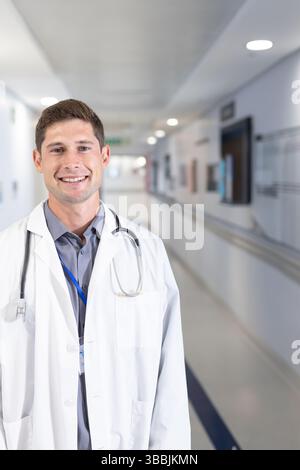 Médecin masculin debout et souriant dans le couloir de l'hôpital, avec stéthoscope et badge d'identification, espace de copie Banque D'Images