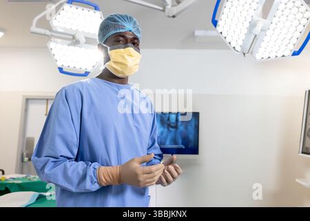 Chirurgien afro-américain portant gants et masque au bloc opératoire avec lumières chirurgicales Banque D'Images
