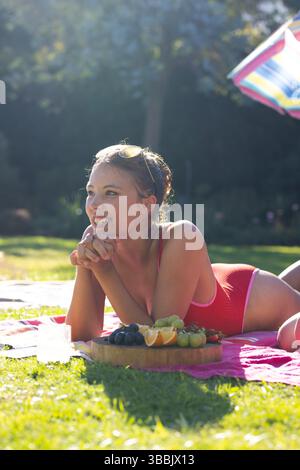 Femme couchée portant maillot de bain rouge sur serviette dans le jardin, avec planche à fruits et boisson sous parapluie Banque D'Images