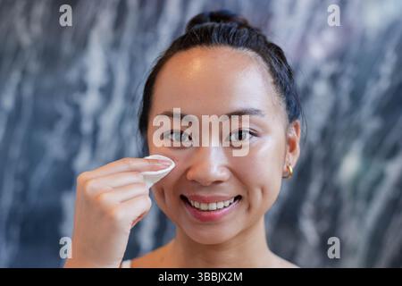Femme asiatique souriante appliquant un tampon de coton à la joue dans la zone de vanité de salle de bains, montrant la routine de soins de la peau Banque D'Images
