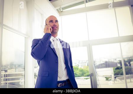Homme d'affaires parlant tenant smartphone portant costume et cravate à motifs près de grandes fenêtres dans le bureau Banque D'Images