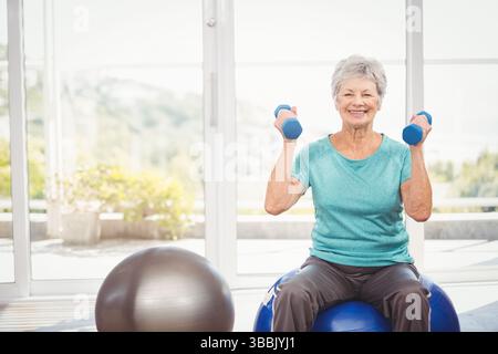 Femme âgée souriante soulevant des haltères bleus dans la salle de gym à domicile, avec boule de stabilité et fenêtres en verre Banque D'Images